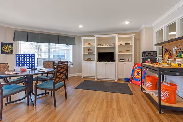 A bright and spacious common room with wood flooring, featuring a round table with four chairs, a dartboard on the wall, a large window with a valance, a white built-in shelving unit with a TV, and a workbench with tools and orange Home Depot buckets.