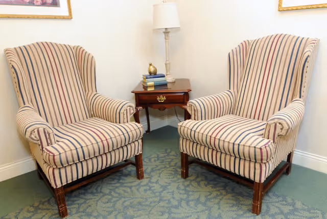 Two striped upholstered armchairs positioned on either side of a wooden side table with a drawer. The table holds a white lamp, a small decorative object, and a stack of books. The setting is indoors with light-colored walls and a patterned carpeted floor.