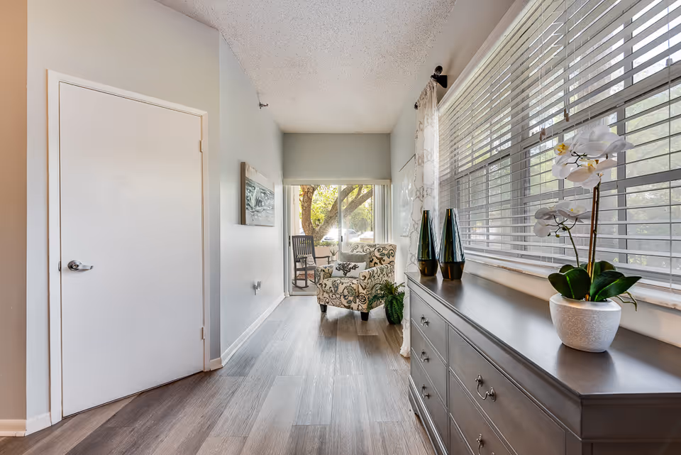 Bright interior hallway leading to a small sitting area with a patterned armchair, dresser, and large window with blinds.