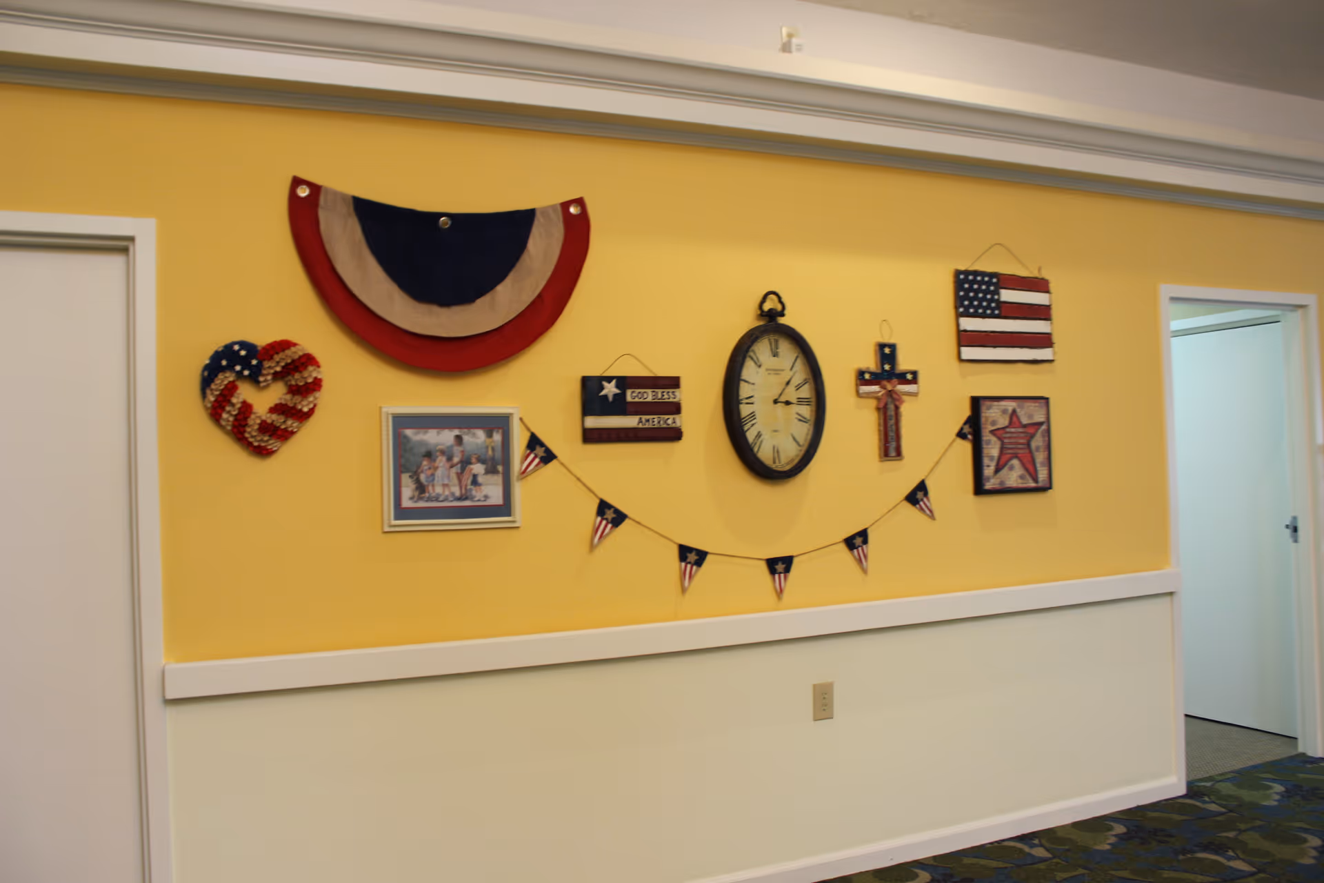 Yellow hallway wall decorated with patriotic wall hangings, a round clock, and a small pennant banner.