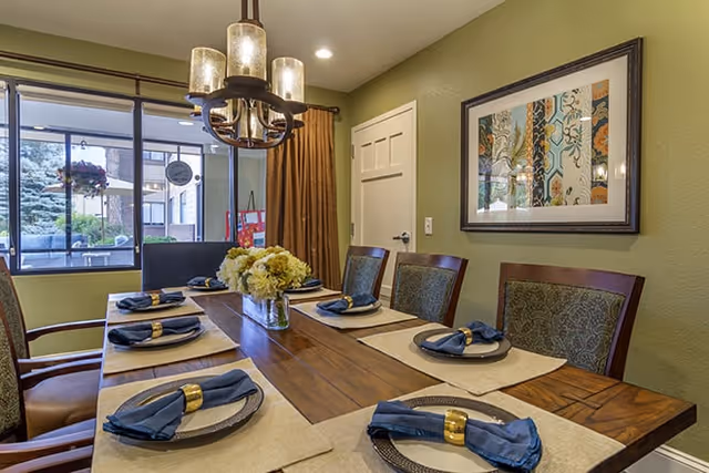 Dining room with a wooden table set for six with blue napkins and gold napkin rings, a floral centerpiece, chandelier, green walls, framed artwork, and sliding glass doors to a patio.