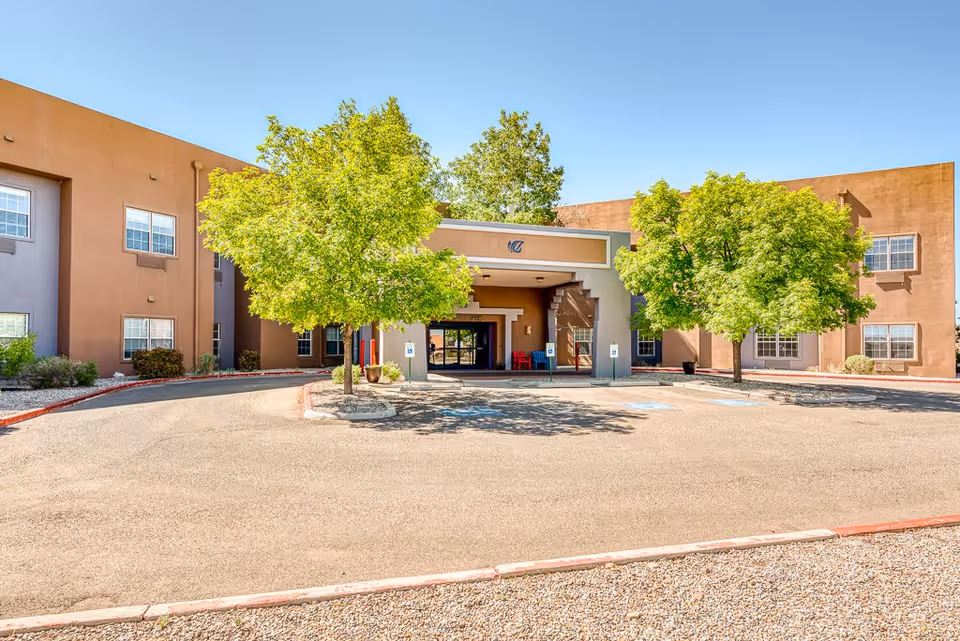 Front exterior view of Sierra Blanca Active Adult Living facility with a driveway, two green leafy trees, and a covered entrance under a clear blue sky.