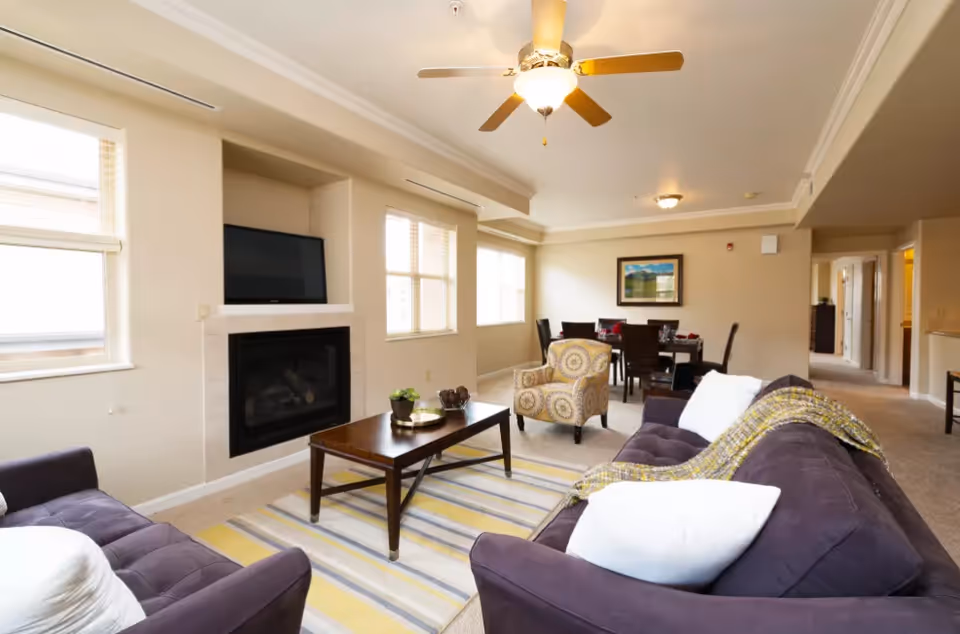 Bright open living room with sofas, a coffee table, fireplace, ceiling fan and a dining area in the background.