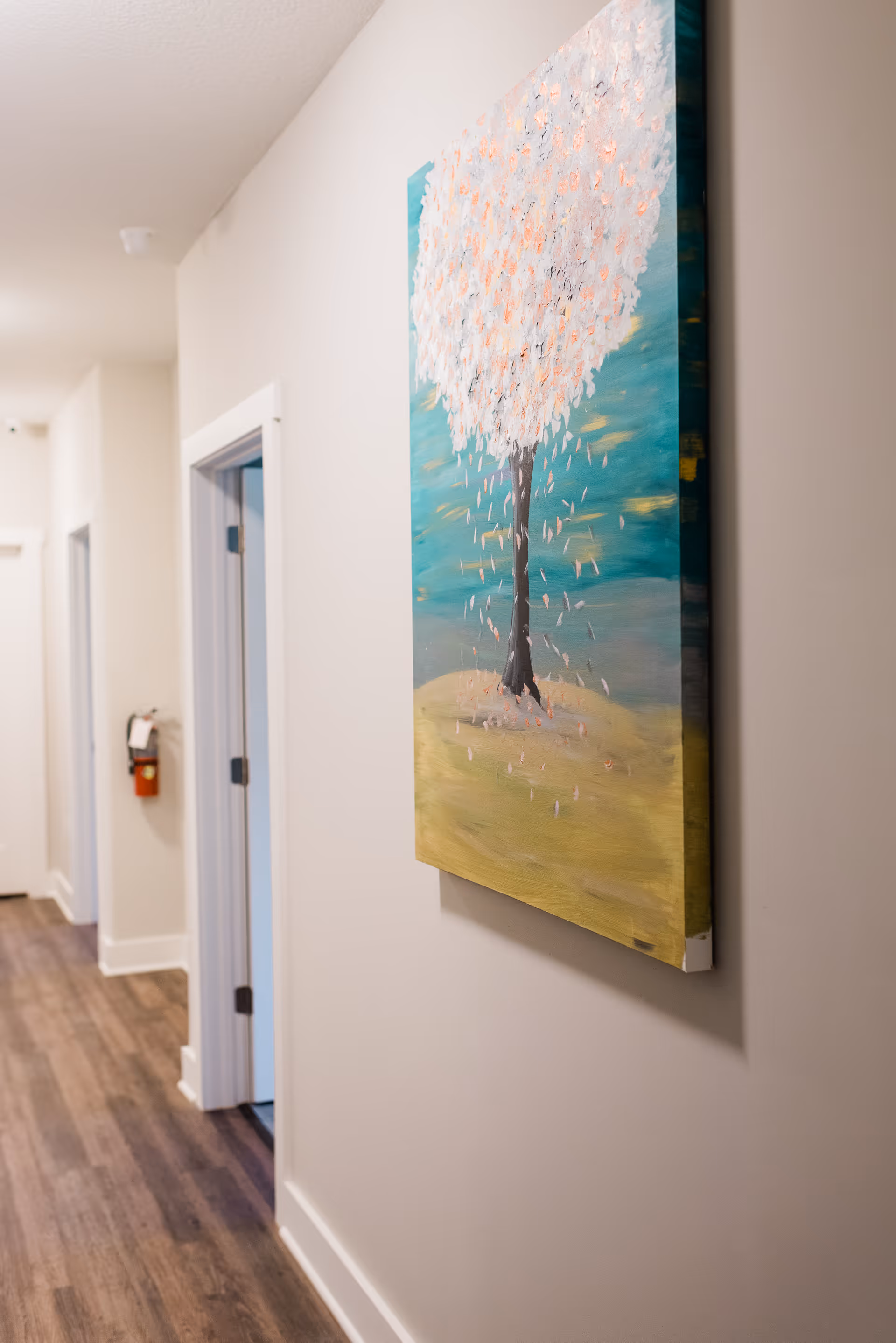 A hallway with wooden flooring and white walls featuring a colorful painting of a tree with white and pink blossoms. There are doorways along the hallway and a fire extinguisher mounted on the wall.