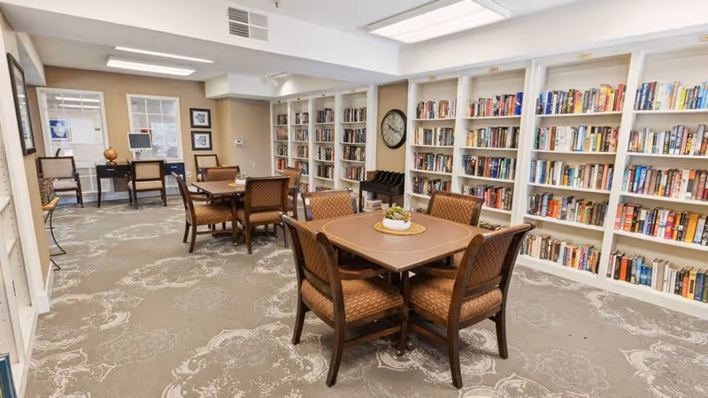 Community library-style common room with wall-to-wall bookshelves and tables surrounded by chairs.