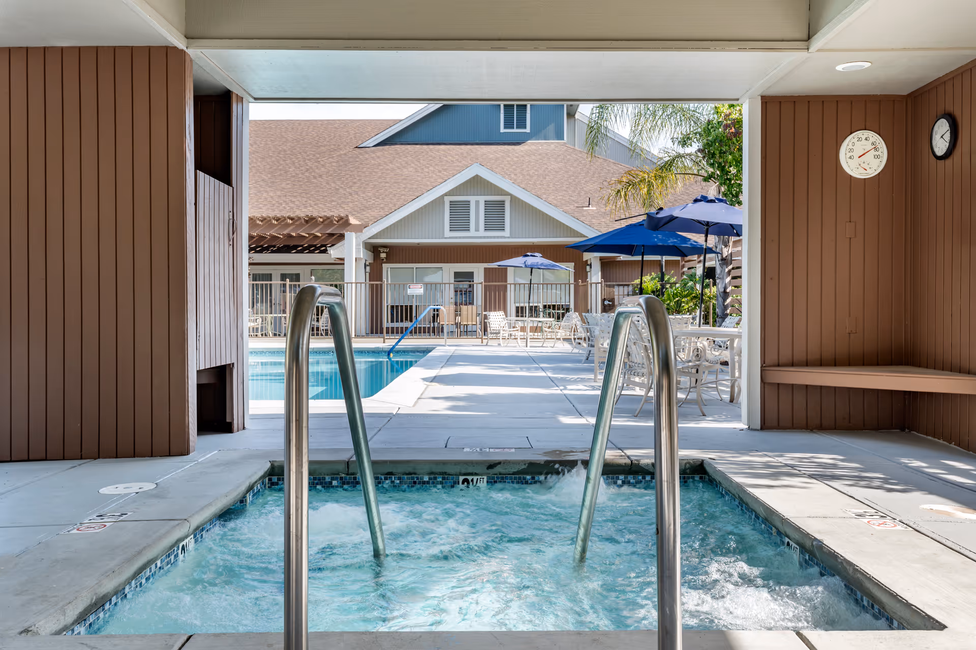 View from inside a covered hot tub area looking out towards an outdoor swimming pool with patio tables, chairs, and blue umbrellas. A building with a brown roof and beige walls is visible in the background. There is a thermometer and a clock mounted on the brown wooden wall to the right.