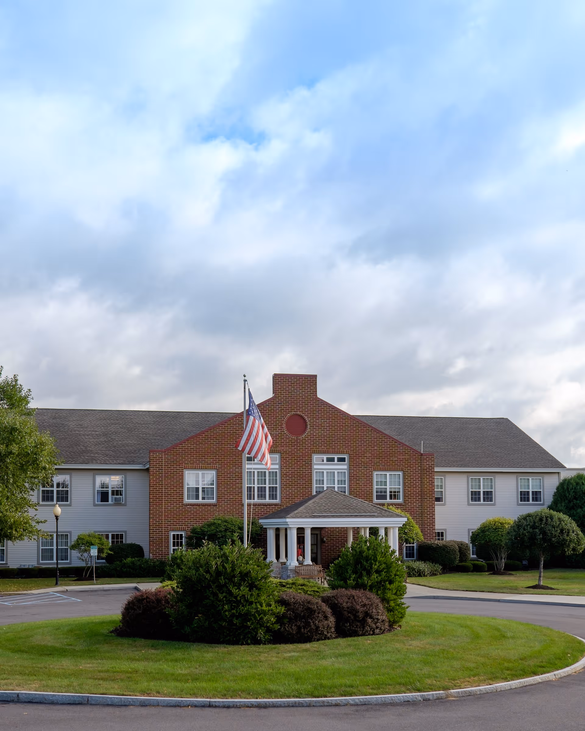 Front exterior view of a two-story senior living facility building with a brick central section and white siding on the wings. There is a circular driveway with a landscaped island featuring bushes and an American flag on a flagpole. The sky is partly cloudy.