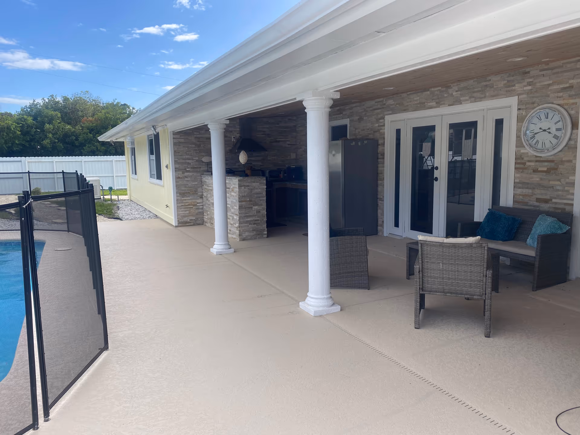 Outdoor covered patio area with stone wall, two white columns, a seating area with wicker chairs and a small sofa with blue cushions, a wall clock, and a kitchen area with a refrigerator and stove. To the left, there is a fenced swimming pool and a white fence enclosing the yard.