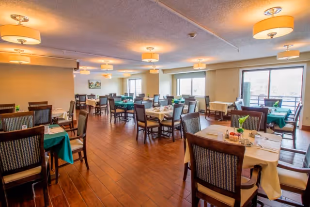 A spacious dining room with multiple tables covered in white and green tablecloths, each set with dining utensils, cups, and small flower arrangements. The room has wooden flooring, large windows letting in natural light, and ceiling lights evenly spaced across the ceiling.