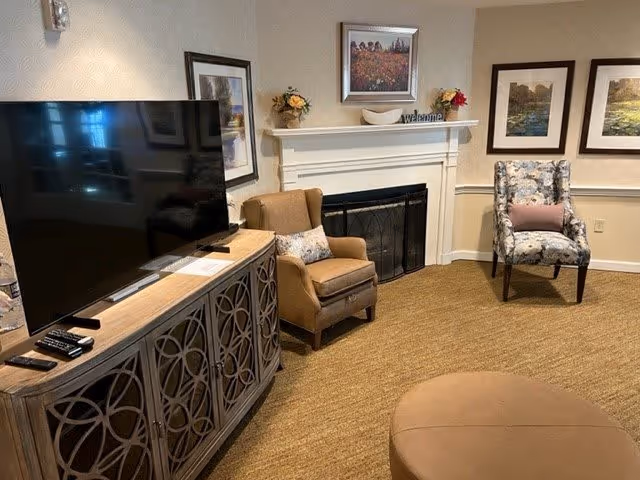 A cozy living room area with a large flat-screen TV on a decorative wooden cabinet, two armchairs—one brown and one floral patterned with a pink pillow, a round ottoman, and a white fireplace with a black screen. Above the fireplace are framed pictures, a small flower arrangement, and a 'welcome' sign.
