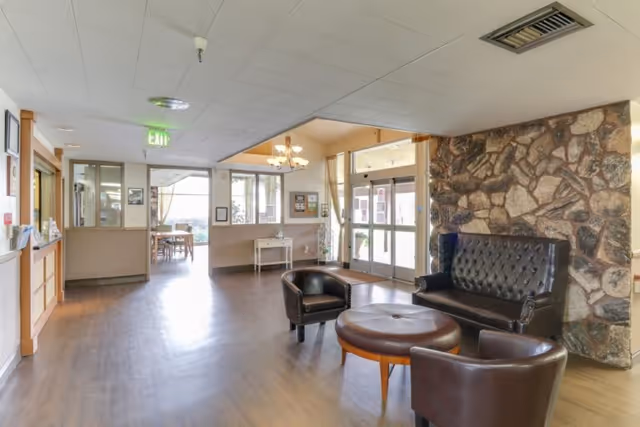 Interior view of a senior living facility lobby area with wooden flooring, a stone accent wall, a black leather sofa, two matching armchairs, and a round ottoman. There is a chandelier hanging from the ceiling near the entrance doors, and a small white table against the wall. Large windows and glass doors allow natural light to fill the space.