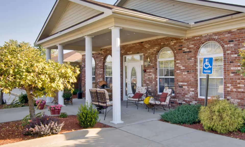 Front entrance of a senior living facility with a covered porch supported by white columns. There are outdoor chairs with cushions and a small table on the porch. The building has a brick exterior with arched windows. A handicapped parking sign is visible near the entrance, and there are landscaped plants and small trees around the walkway.