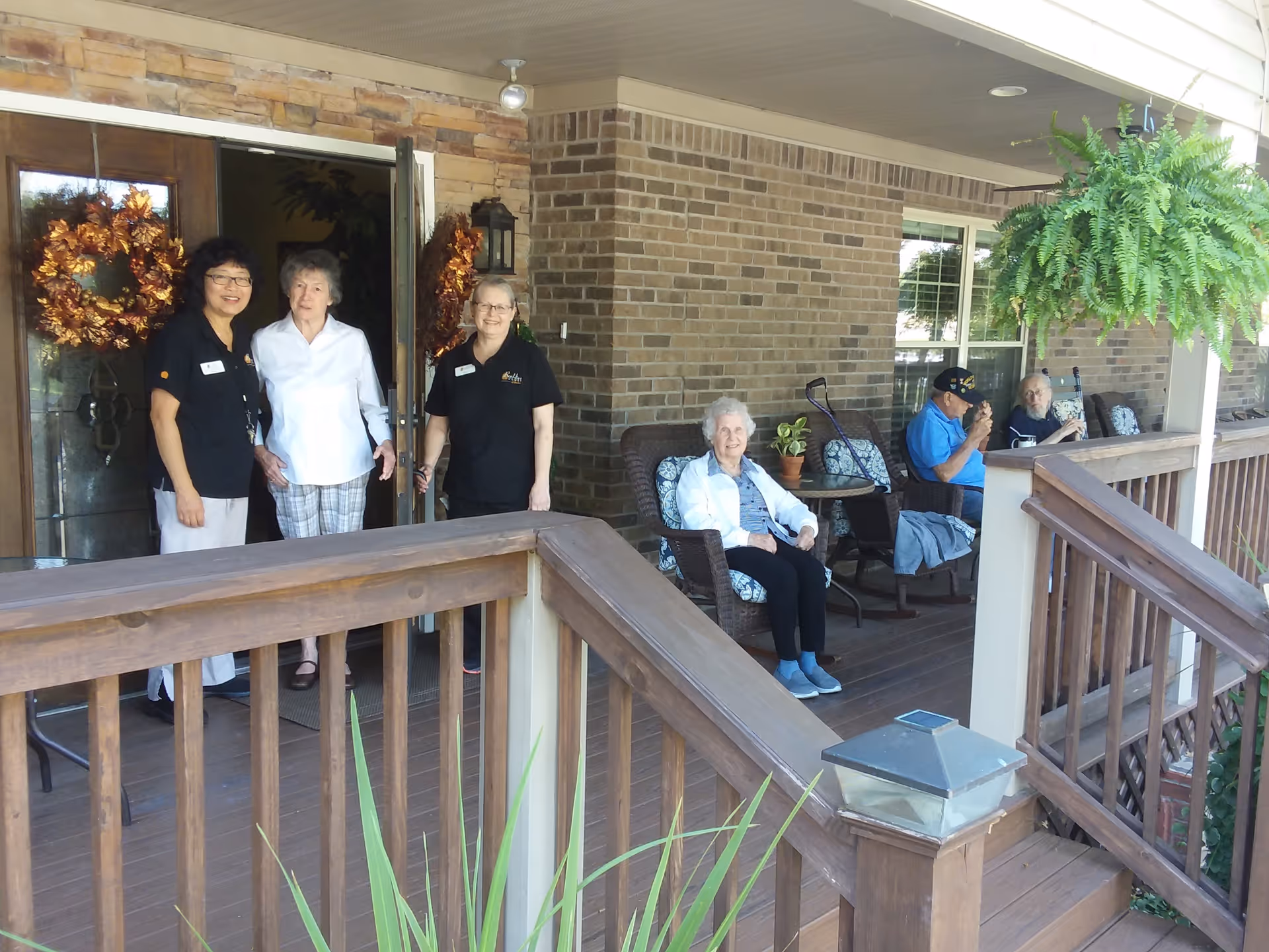 A group of three women standing near the entrance door of a brick building, with two of them wearing black shirts and name tags, and one elderly woman in white. On the porch, three elderly people are seated on cushioned chairs, with a wooden railing and a hanging fern plant visible.