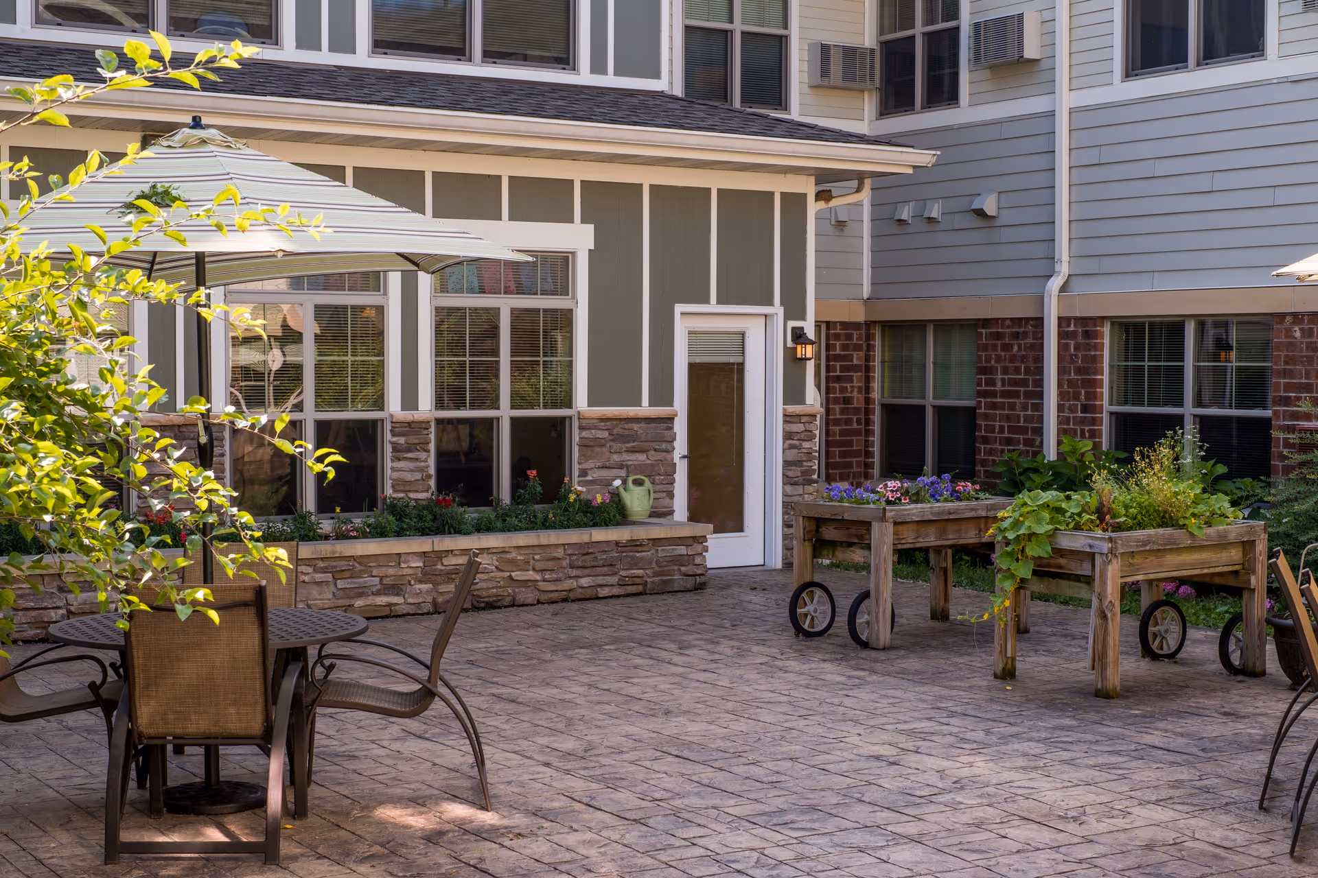 Sunny courtyard patio with tables and chairs under an umbrella, raised planter beds on wheels, and the building facade with windows and a door.