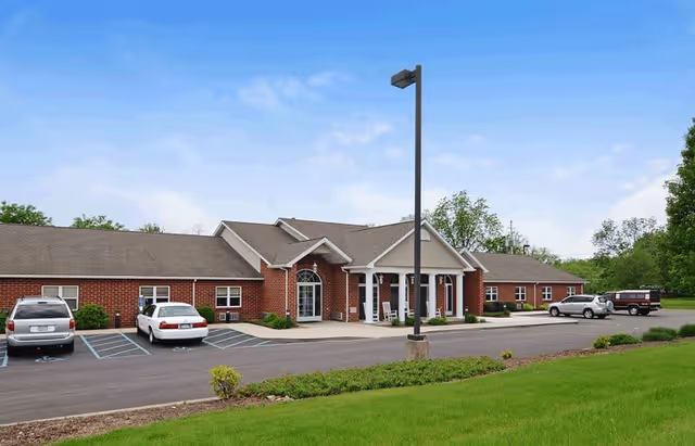 Exterior view of a single-story brick building with a covered entrance supported by white columns. Several cars are parked in front of the building in designated parking spaces. The sky is clear with a few clouds, and there is green grass and some small shrubs in the foreground.