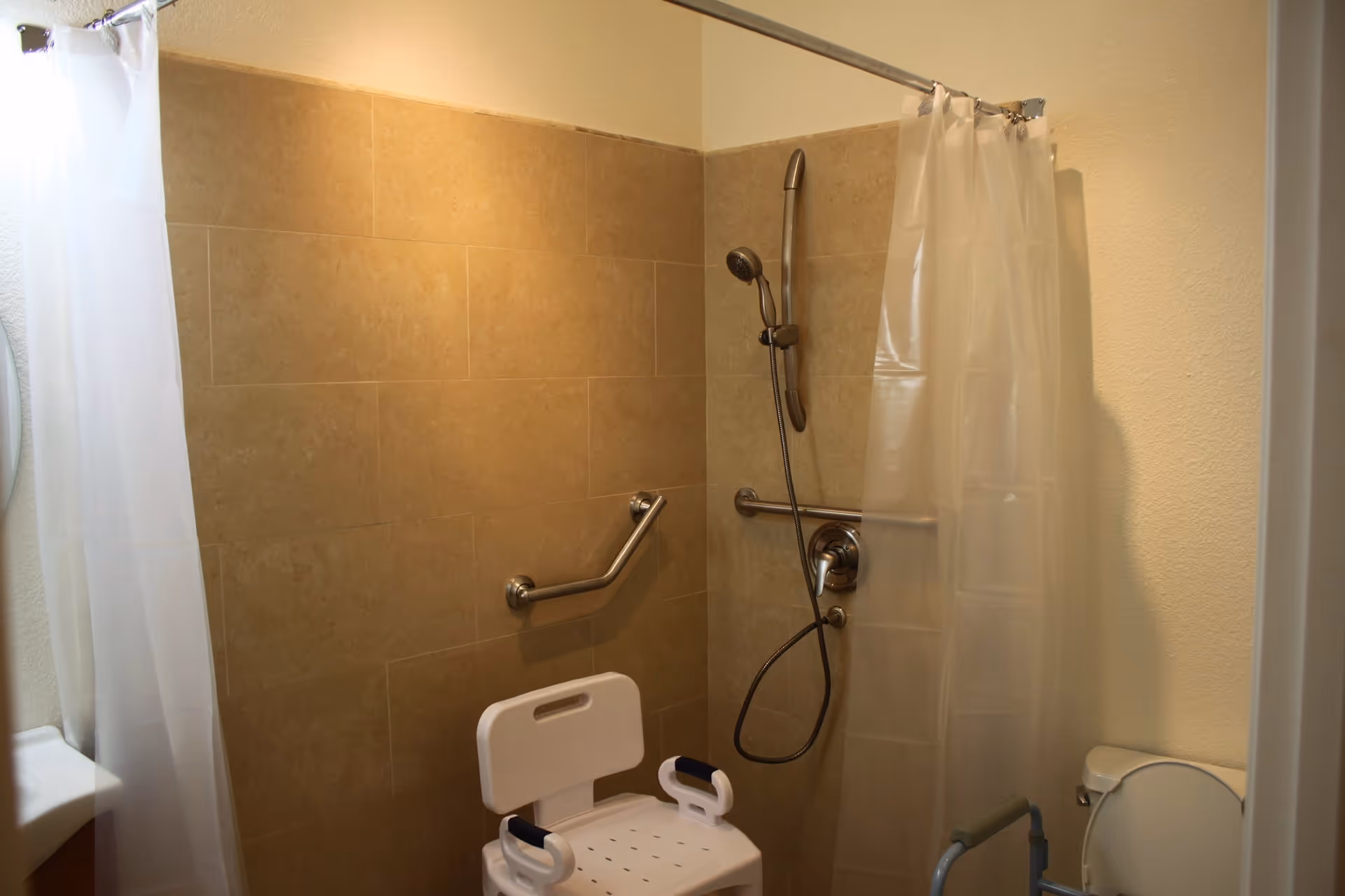 A bathroom with beige tiled walls, a white shower chair with armrests, a handheld showerhead attached to a metal bar, grab bars on the wall, a white shower curtain partially drawn, and a toilet with a support rail next to it.