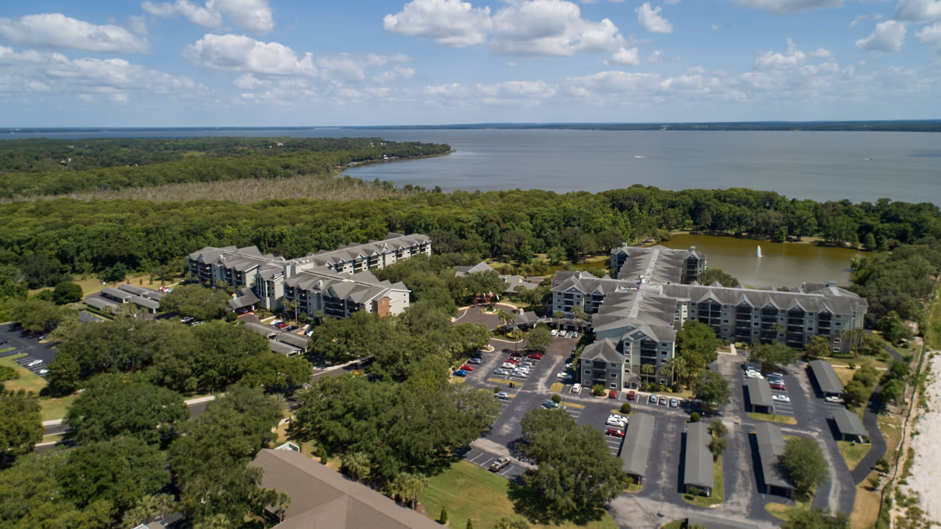 Aerial view of the Lake Port Square complex with multiple apartment buildings, parking areas, trees, and a large lake in the background.
