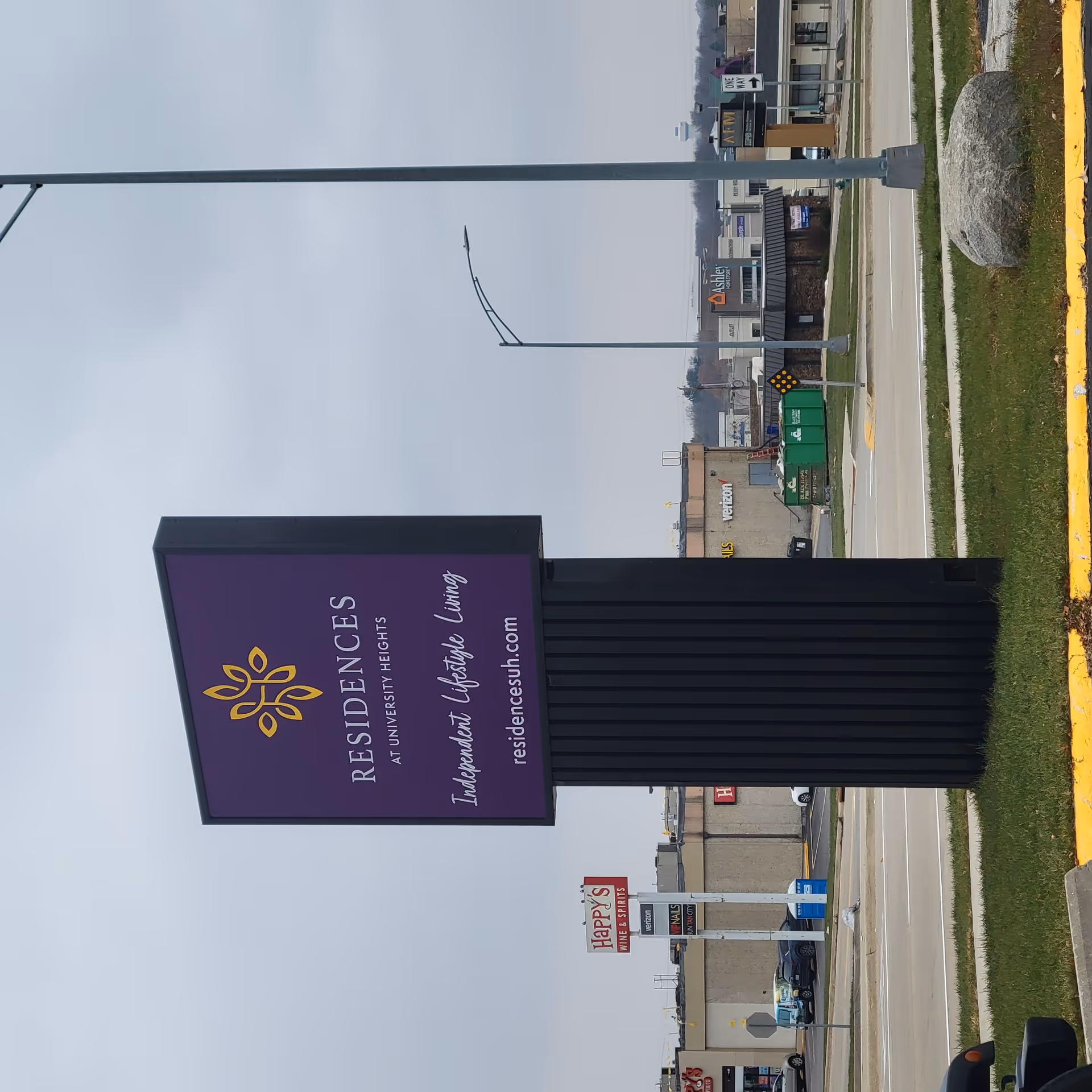 Purple freestanding sign for "Residences at University Heights" beside a road with a shopping center in the background.