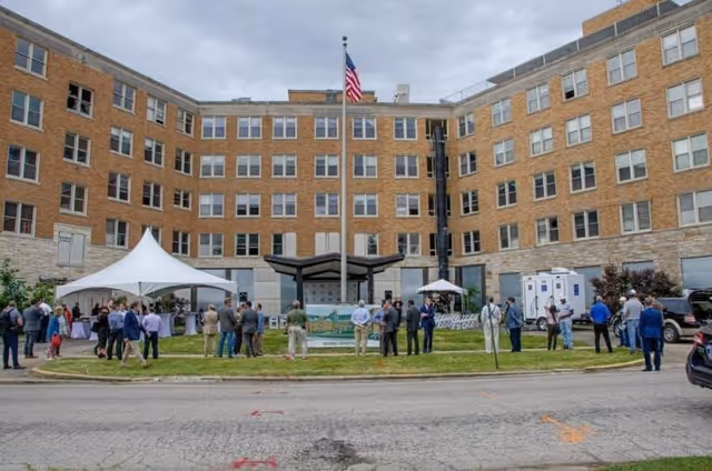 A group of people gathered outside a large multi-story brick building with many windows. There is a flagpole with an American flag in the center of a grassy area, and two white tents set up on the lawn. The scene appears to be an outdoor event or gathering in front of the building.