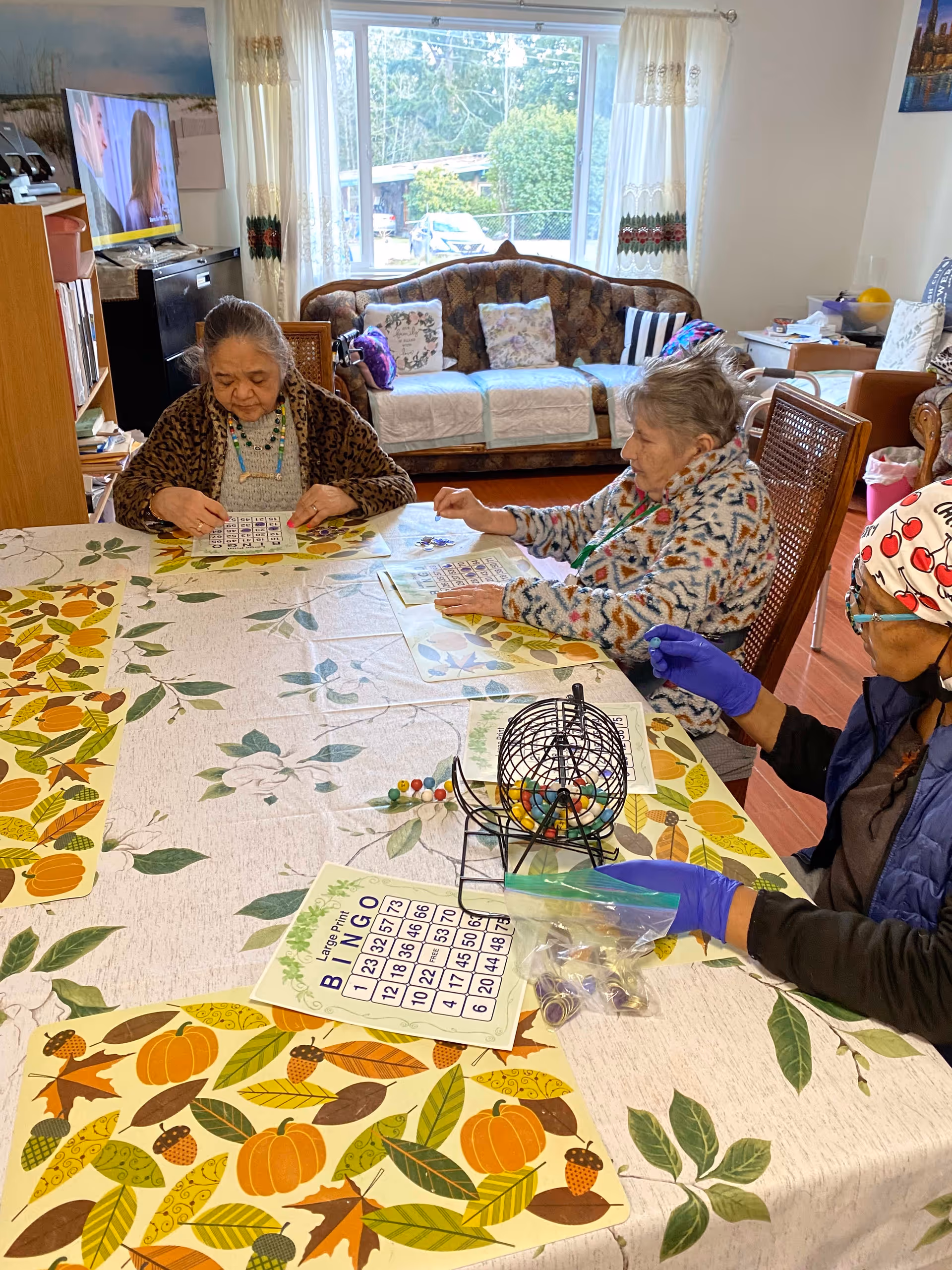 Three elderly women sit around a table playing bingo in a communal living/dining area with a couch and large window in the background.