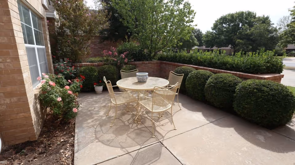 Small outdoor patio with a round metal table and four chairs surrounded by trimmed bushes and flowering plants next to a brick building.