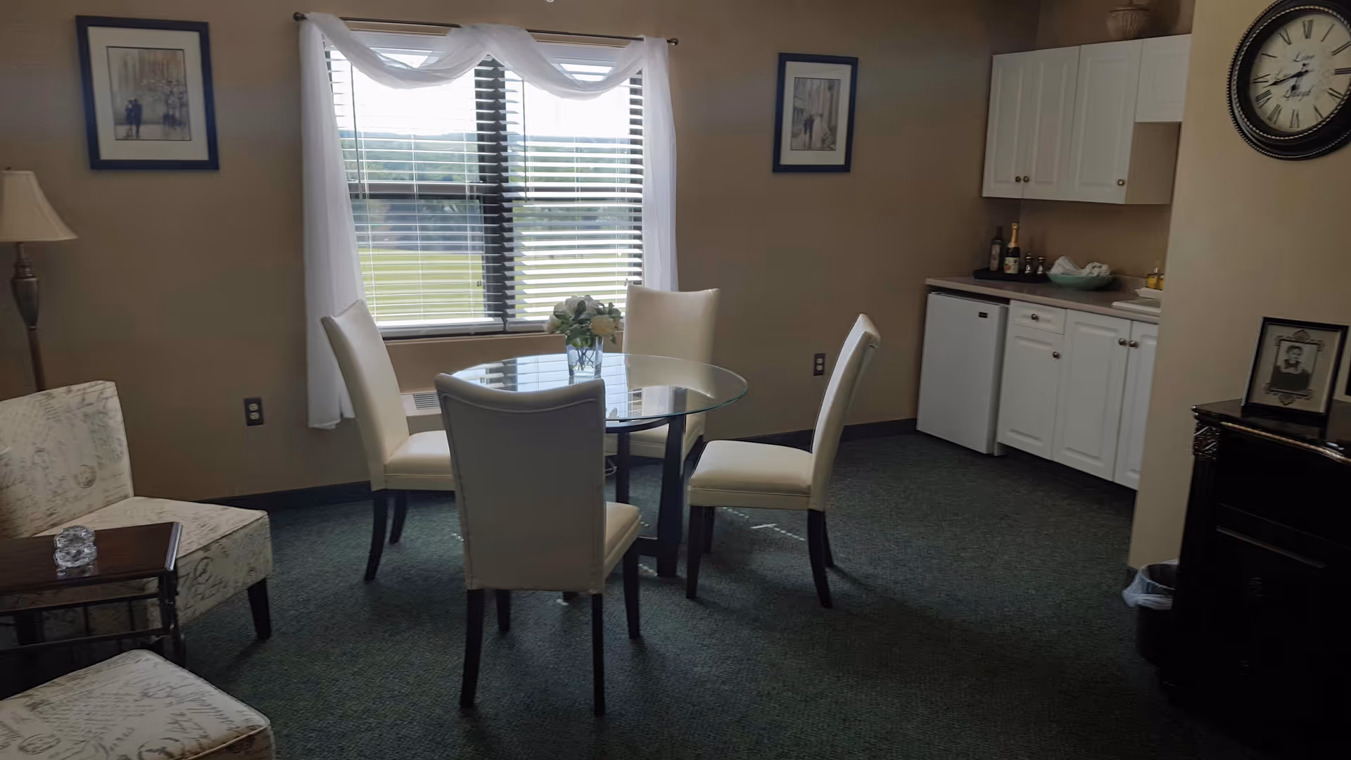 A small dining area with a round glass table surrounded by four white upholstered chairs. There is a window with white curtains letting in natural light. To the right, there is a kitchenette with white cabinets, a mini fridge, and a countertop with some bottles and a bowl. The room has green carpet and beige walls, decorated with framed pictures and a clock.
