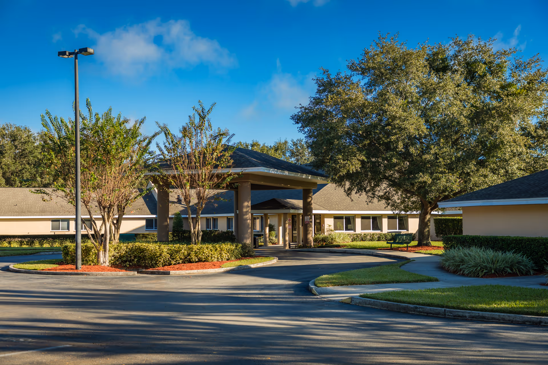 Front entrance of a single-story senior living facility with a covered porte-cochère, landscaped beds, and large trees under a blue sky.