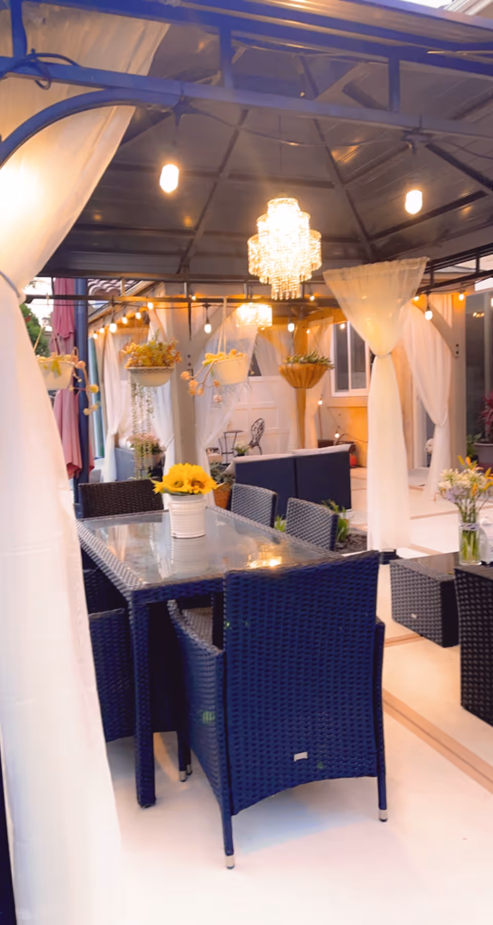 Outdoor covered patio area with a glass-top dining table surrounded by dark wicker chairs. The space is decorated with hanging flower pots, white sheer curtains, and warm lighting including a chandelier and string lights. There are additional seating areas with dark wicker furniture and flower arrangements.