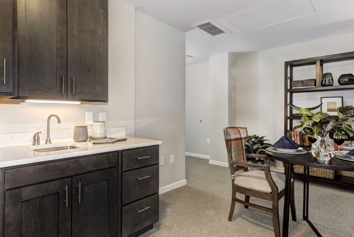 A kitchenette with dark wood cabinets and a small dining table and chairs beside a shelving unit in a senior living interior.