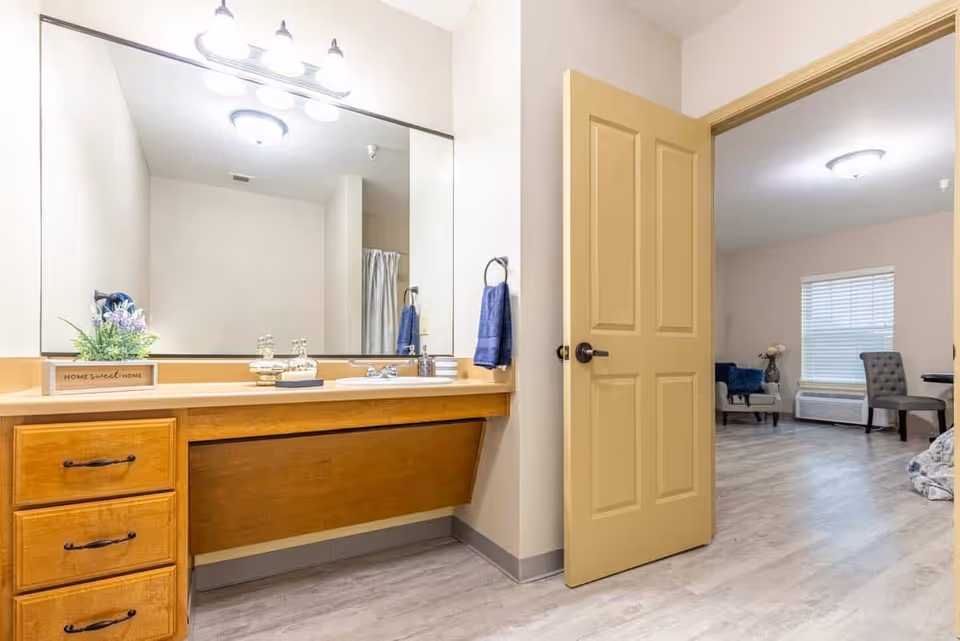 A bathroom vanity area with a large mirror above a wooden countertop and drawers. There is a sink with a faucet, a small plant in a wooden box labeled 'Home Sweet Home', and decorative items on the counter. A blue towel hangs on a ring on the wall next to an open door leading to a room with chairs, a window with blinds, and a light fixture on the ceiling.