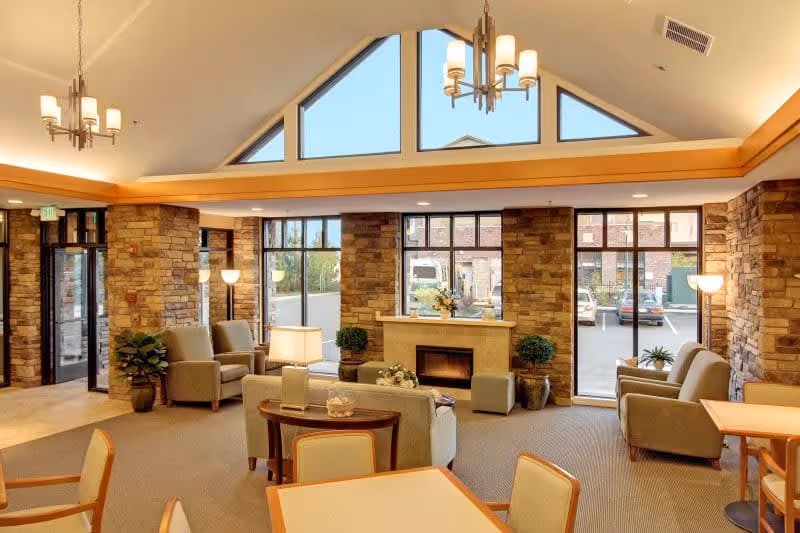 A spacious and well-lit senior living facility common area with large windows, stone accent walls, and a fireplace. The room is furnished with comfortable armchairs, sofas, tables, and lamps, creating a cozy and inviting atmosphere. Outside the windows, a parking lot and some buildings are visible.
