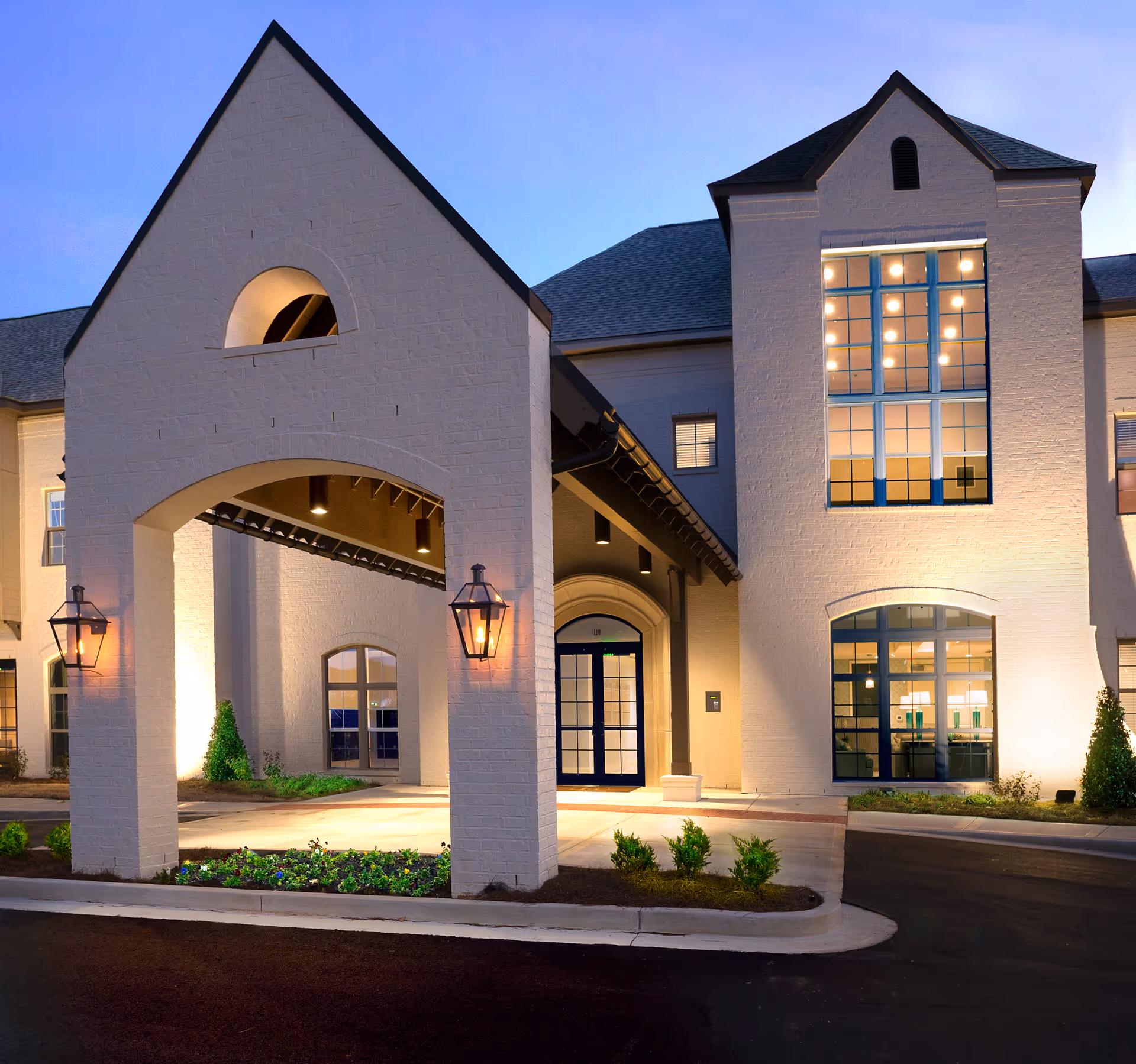 Exterior view of The Magnolia at Oxford Commons building entrance at dusk, featuring a covered driveway with lantern-style lights and large windows illuminated from inside.