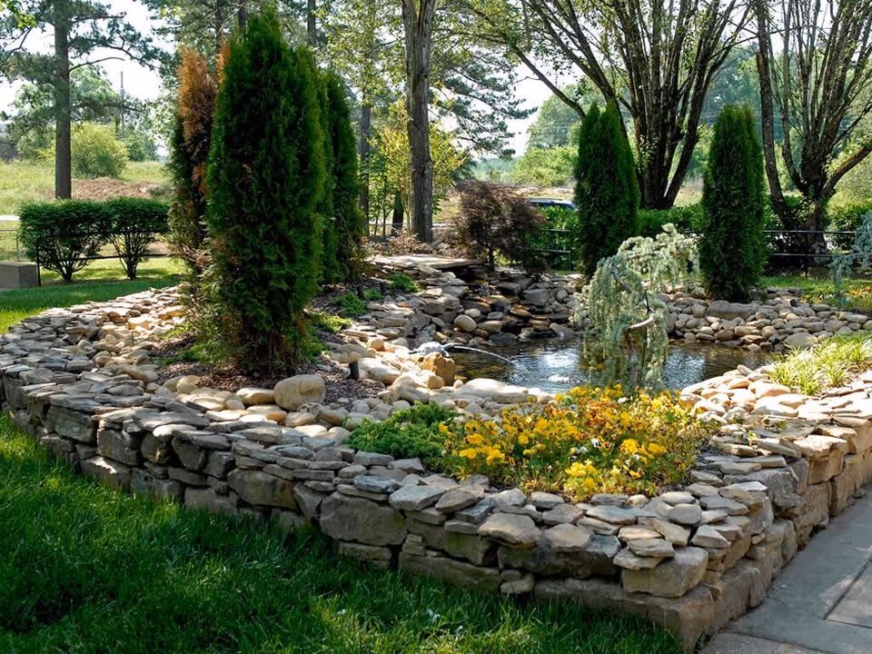 A landscaped outdoor garden area featuring a stone-bordered pond with a small waterfall, surrounded by various green shrubs, trees, and yellow flowers under a sunny sky.