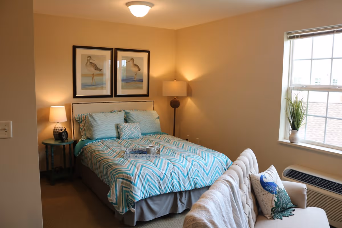 A cozy bedroom with a bed covered in a blue and white chevron-patterned comforter and matching pillows. There are two framed bird pictures hanging above the bed. A small round side table with a lamp and an alarm clock is on one side of the bed, and a floor lamp is in the corner. A window with blinds lets in natural light, and a small potted plant sits on the windowsill. In front of the bed is a cushioned bench with a decorative pillow and a blanket draped over it.
