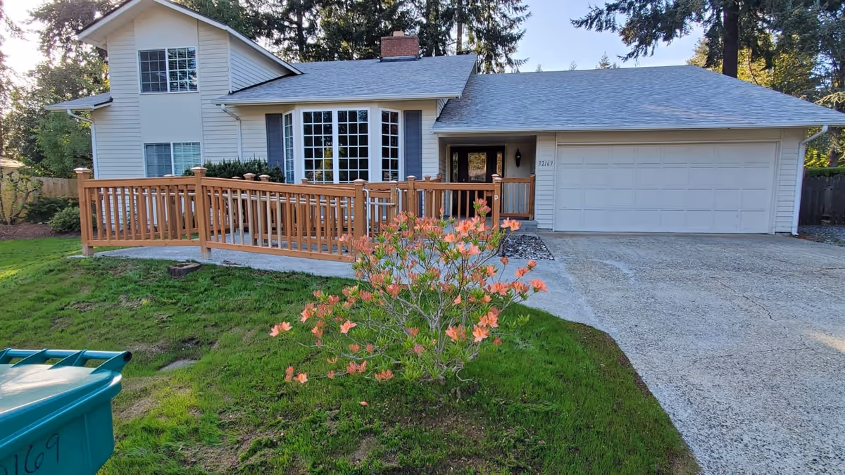 Front exterior view of a two-story house with white siding and a gray roof. There is a wooden ramp leading to the front door, a large bay window, and a two-car garage. A flowering shrub with orange blossoms is in the green lawn in front of the house. Trees and other greenery surround the property.