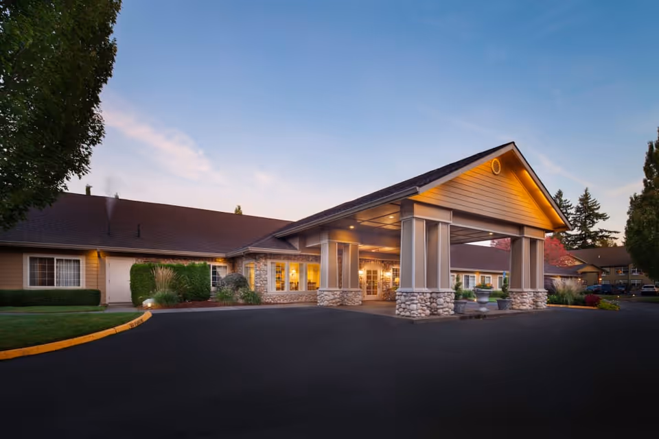 Exterior view of Cedar Crest Alzheimer's Special Care Center at dusk, showing a single-story building with a covered entrance supported by stone and pillar columns, surrounded by landscaped greenery and a paved driveway.