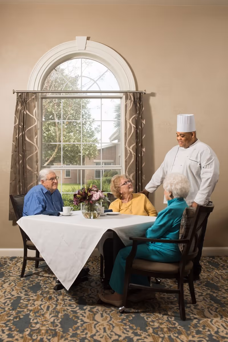 Three elderly people sitting around a dining table with a white tablecloth and a vase of flowers, interacting with a chef in a white uniform and hat in a room with a large arched window and patterned curtains.