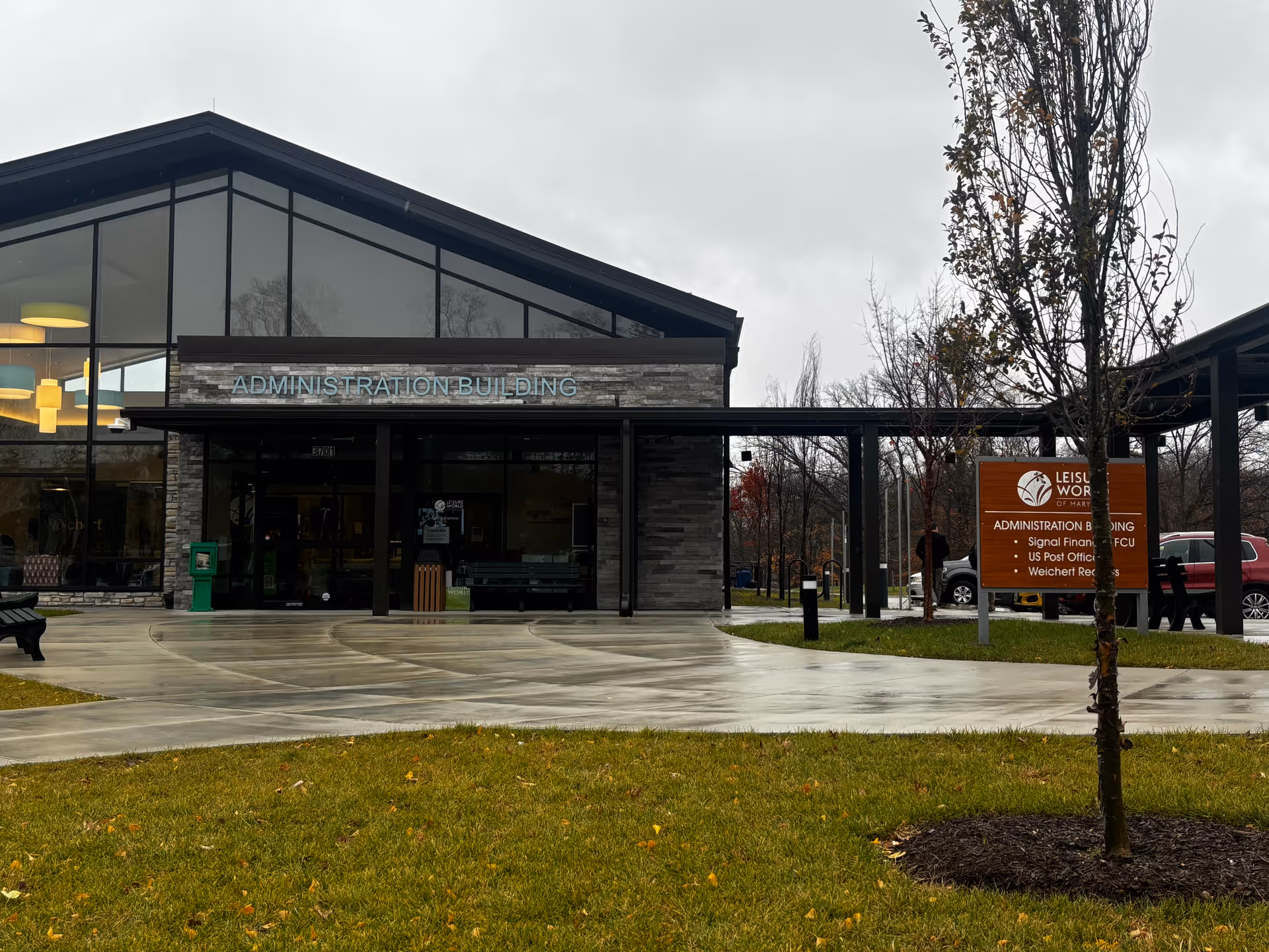 Exterior view of the Leisure World of Maryland Administration Building on a cloudy day, showing a modern building with large glass windows, a paved walkway, green grass, and a sign listing services including Signal Financial FCU, US Post Office, and Weichert Realtors.