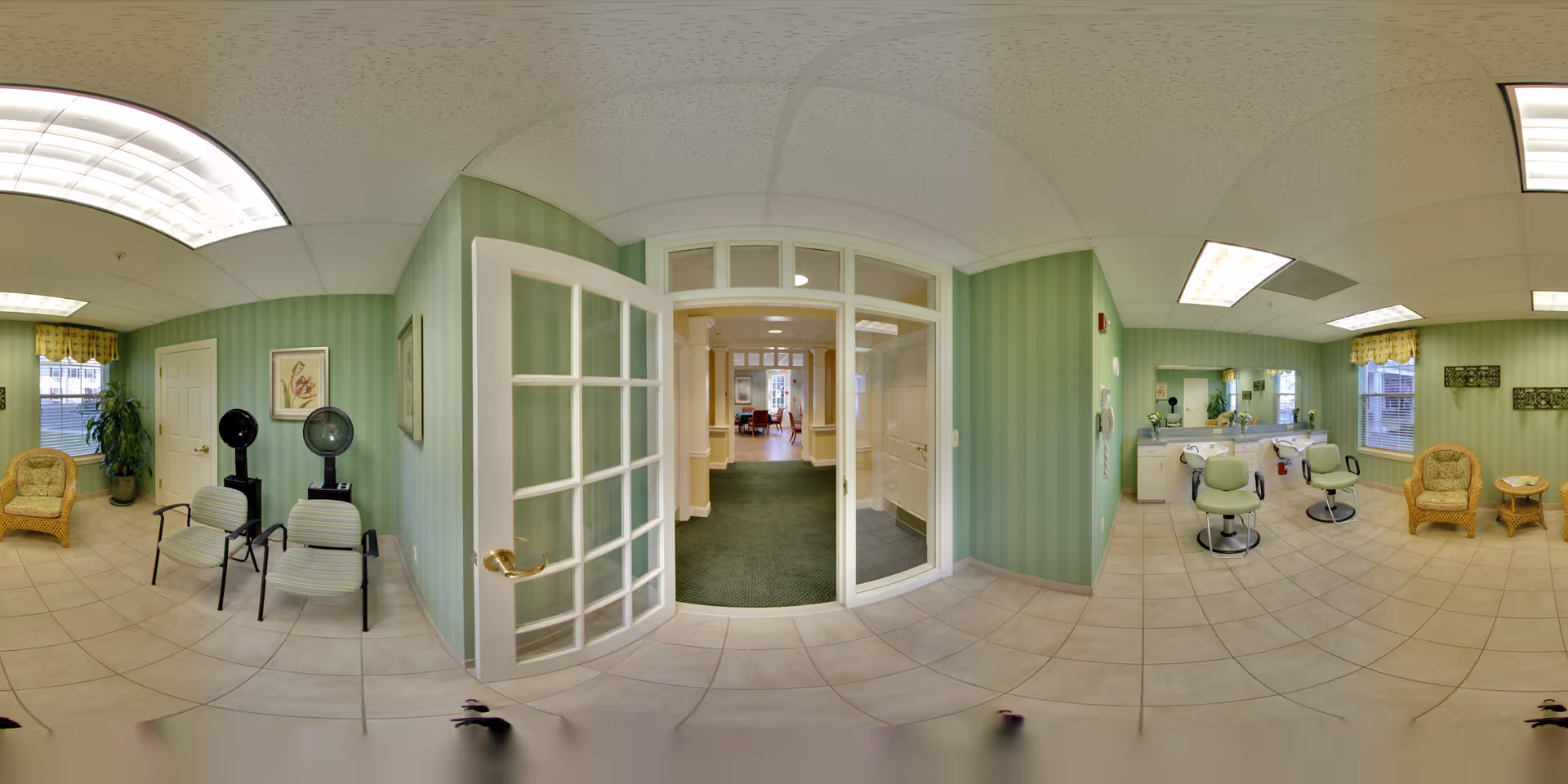 Interior view of a senior living facility's salon area with green striped walls and tiled floor. The room features two hair drying chairs with hooded dryers, two salon chairs in front of mirrors and sinks, wicker chairs with a small table, and a potted plant near a window. A glass-paned door opens to a hallway with carpeted flooring and additional rooms visible in the distance.