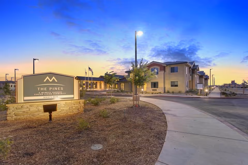 Exterior front entrance of The Pines senior living community at dusk with a lit sign, curved driveway, and the building in the background.