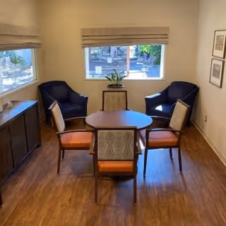 A small sitting area with a round wooden table surrounded by four chairs, two with orange cushions and two with patterned backs. There are two dark blue armchairs positioned near a window with beige blinds. A wooden cabinet is placed against the left wall, and two framed pictures hang on the right wall. The floor is wooden, and natural light comes through the windows.