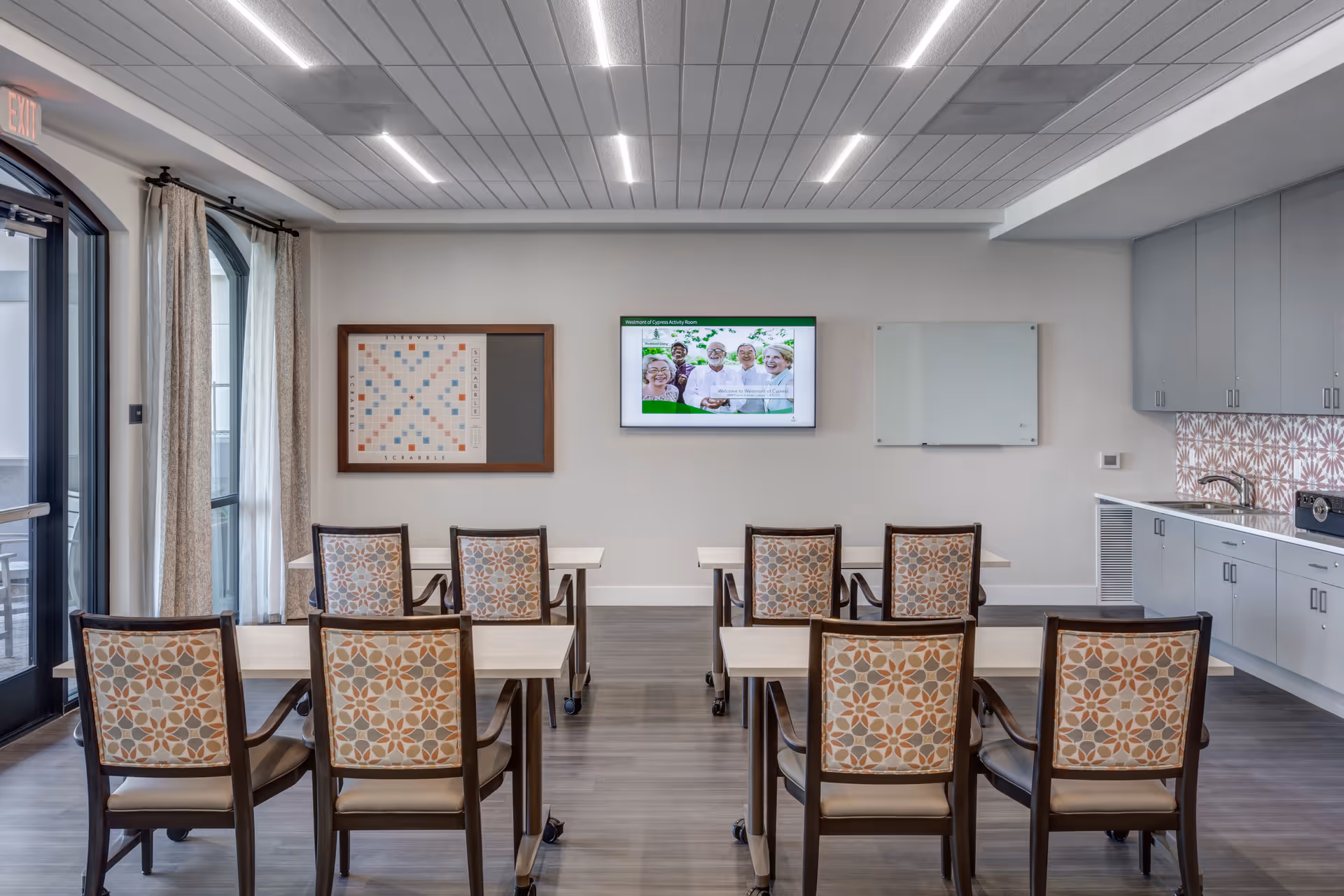 Well-lit activity room with rows of tables and patterned chairs facing a wall-mounted TV, a game board, whiteboard, and a kitchenette.