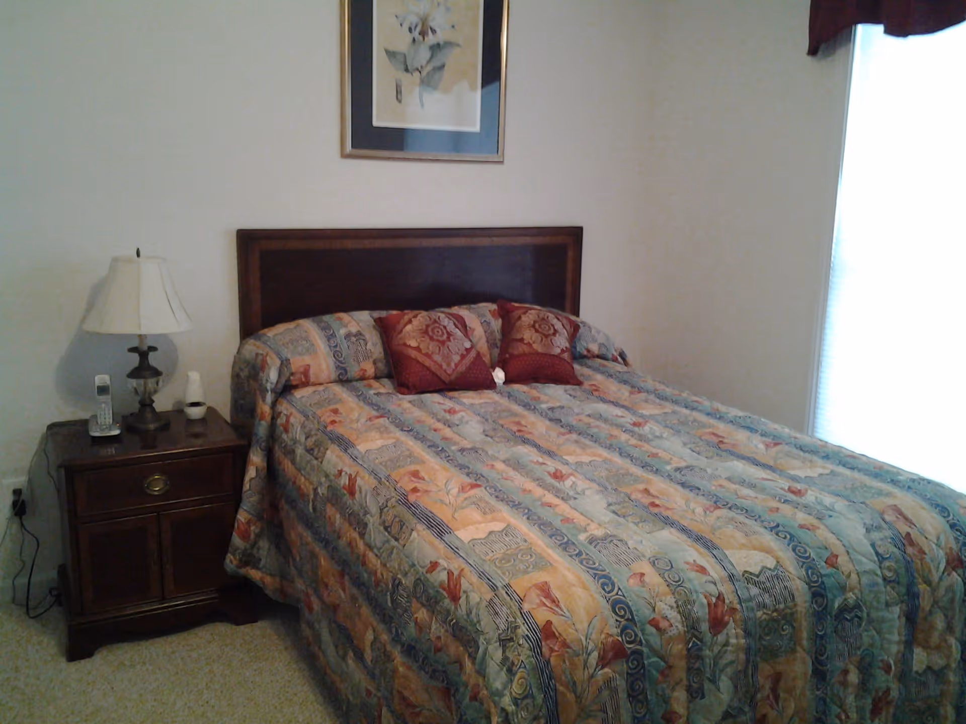 A bedroom with a bed covered in a colorful patterned bedspread and two decorative pillows. There is a wooden nightstand with a lamp, a cordless phone, and a small item on it. A framed floral picture hangs on the wall above the bed, and a window with blinds is partially visible on the right side.