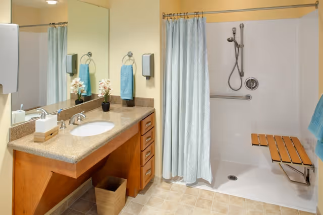A clean and accessible bathroom featuring a wooden vanity with a granite countertop, an oval sink, a large mirror, and a small potted plant. There is a mounted soap dispenser and a blue hand towel hanging on a ring. The walk-in shower has a white tiled wall, a handheld showerhead, a grab bar, and a fold-down wooden shower seat. The shower curtain is light blue with a subtle pattern.