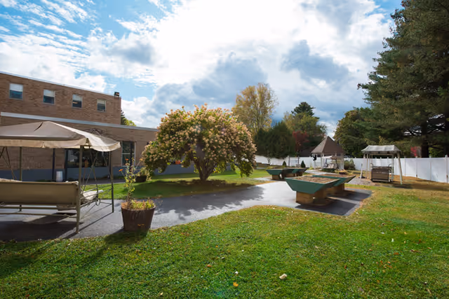 Outdoor courtyard with a grassy lawn, flowering tree, covered swings and picnic tables next to a low brick building under a partly cloudy sky.