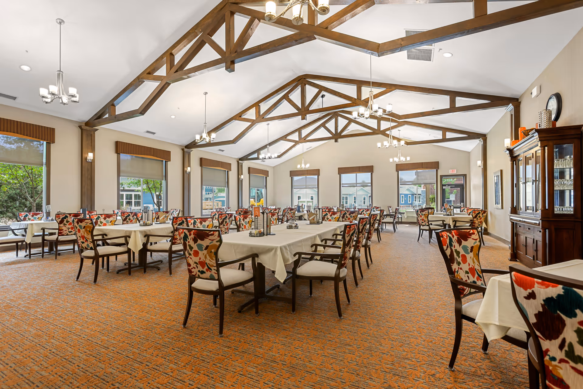 A spacious dining room with multiple tables covered with white tablecloths and surrounded by chairs with colorful floral upholstery. The room features large windows with brown valances, wooden ceiling beams, and several chandeliers providing warm lighting. A wooden cabinet with glassware is visible on the right side of the room.