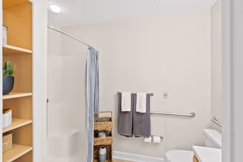 Bright accessible bathroom with a shower and curtain, grab bar, toilet, wicker storage and towels on a rail.