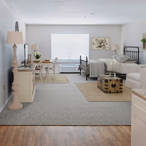 A bright and spacious studio apartment featuring a bed with patterned bedding, a small dining table with chairs, a TV on a white stand, a beige armchair, a wooden coffee table, and a large window with blinds letting in natural light. The room has light-colored walls and a mix of carpet and wood flooring.