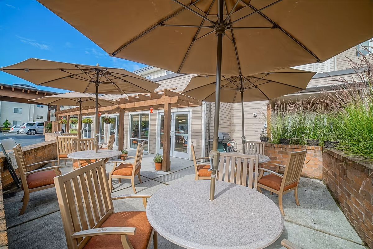 Outdoor patio area with round tables and wooden chairs with orange cushions, shaded by large beige umbrellas. The patio is adjacent to a building with glass doors and windows, surrounded by brick planters with tall grasses and hanging plants. A clear blue sky is visible above.