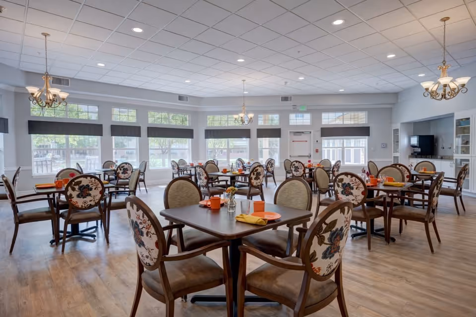 A bright and spacious dining room with multiple tables and chairs arranged neatly. Each table is set with orange plates, cups, and yellow napkins. The chairs have floral patterned upholstery on the backrests. Large windows with black valances allow natural light to fill the room. Two chandeliers hang from the ceiling, and there is a beverage station with a coffee machine on the right side of the room.